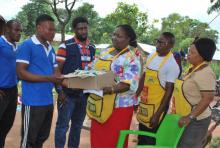 State Technical Facilitator; Ogoja LGA Mrs Ebom Okoma presenting relief materials to the Chairman of the 41 communities in Adagom Refugee Camp; Elias Taku alongside other government officials in yellow aprons.